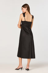 A woman is wearing a black satin sleeveless dress with lace and ribbon details on the shoulder straps. The dress has a fitted bodice and flows down to a midi length. She is also wearing black open-toe heeled sandals.