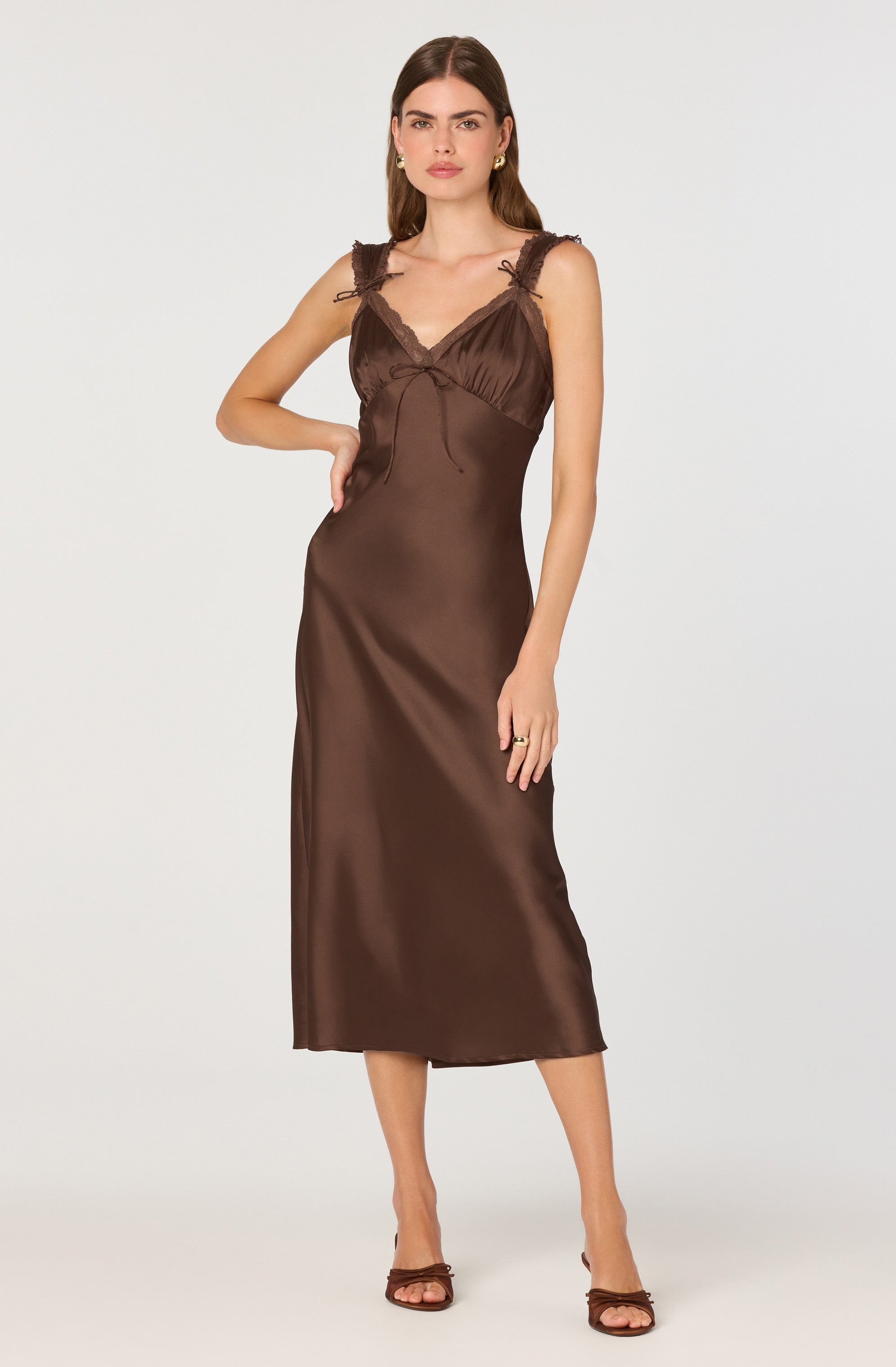 A woman is wearing a sleeveless, brown satin dress that features lace detailing around the neckline and shoulder straps. The dress has a fitted bodice with a small bow at the center and flows down to a longer length, ending mid-calf. She is also wearing matching brown high-heeled open-toe sandals.