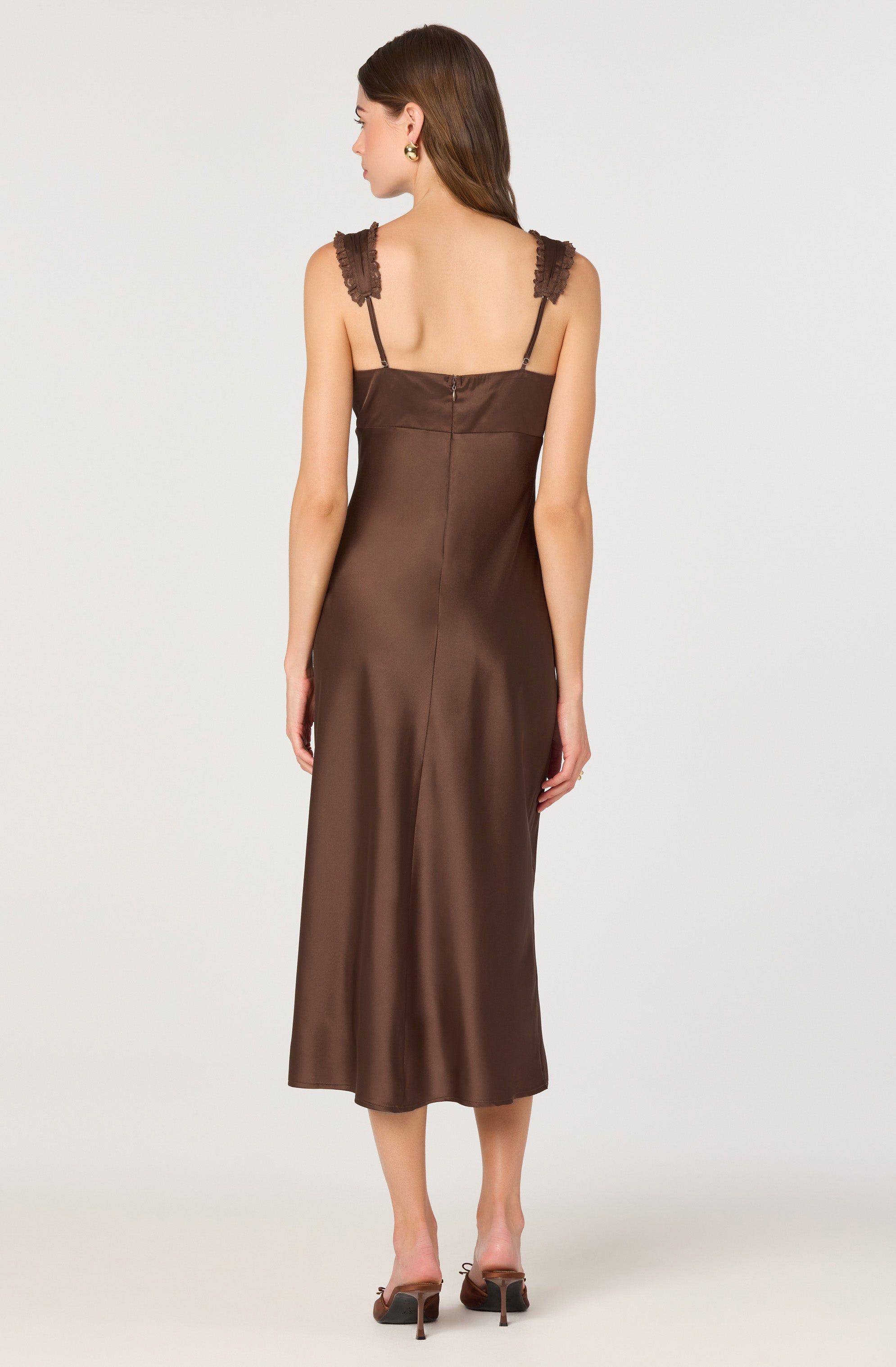 A woman is wearing a long, brown satin slip dress with lace shoulder straps. The dress has a fitted bodice, a back zipper closure, and a flowy, ankle-length skirt. She is also wearing high-heeled sandals.