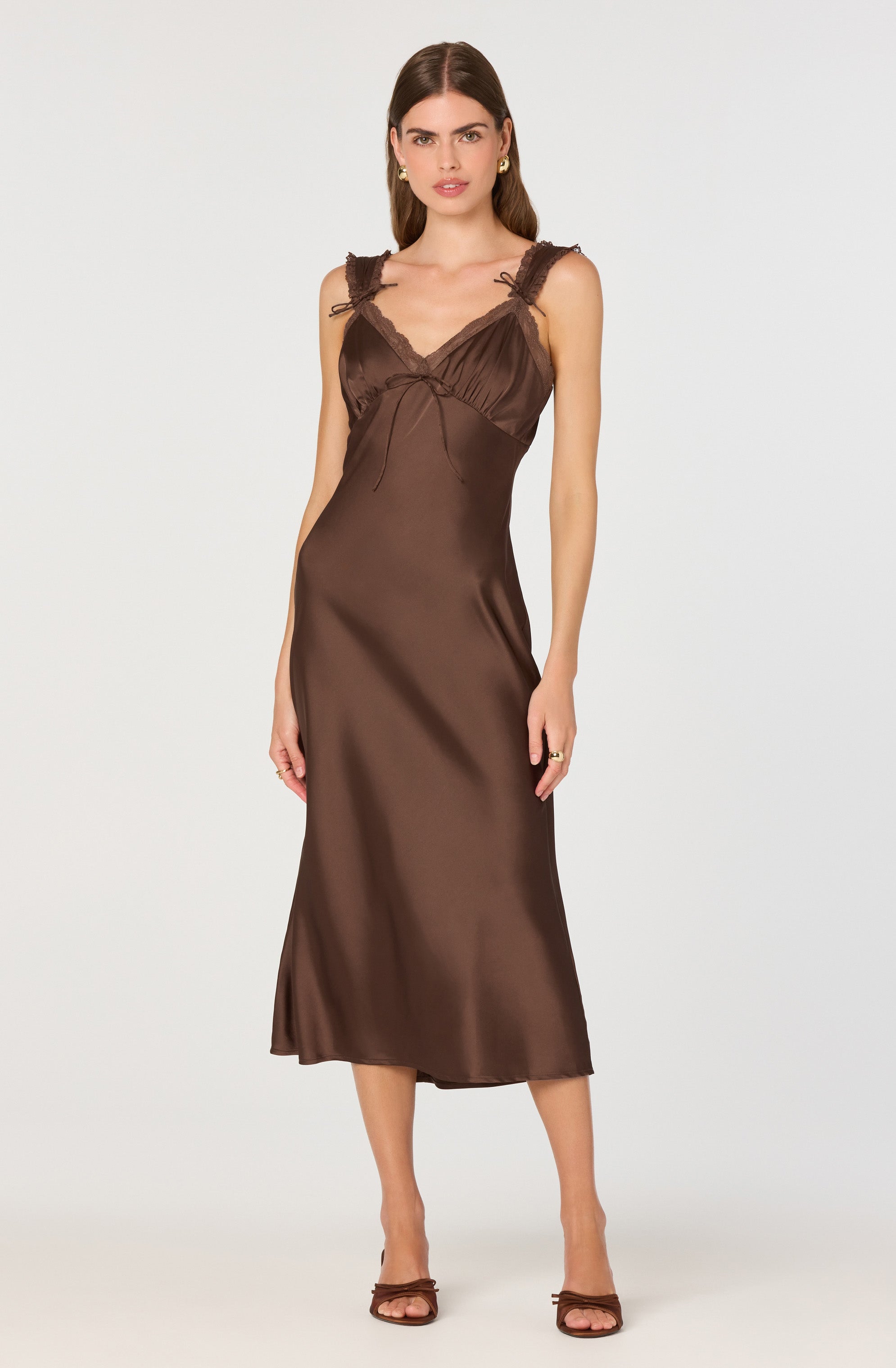 A woman is wearing a long, brown satin slip dress that reaches mid-calf. The dress features ruffled straps with lace detailing and a V-neckline with subtle ruching at the bust. She is also wearing dark brown, open-toe high-heeled sandals.