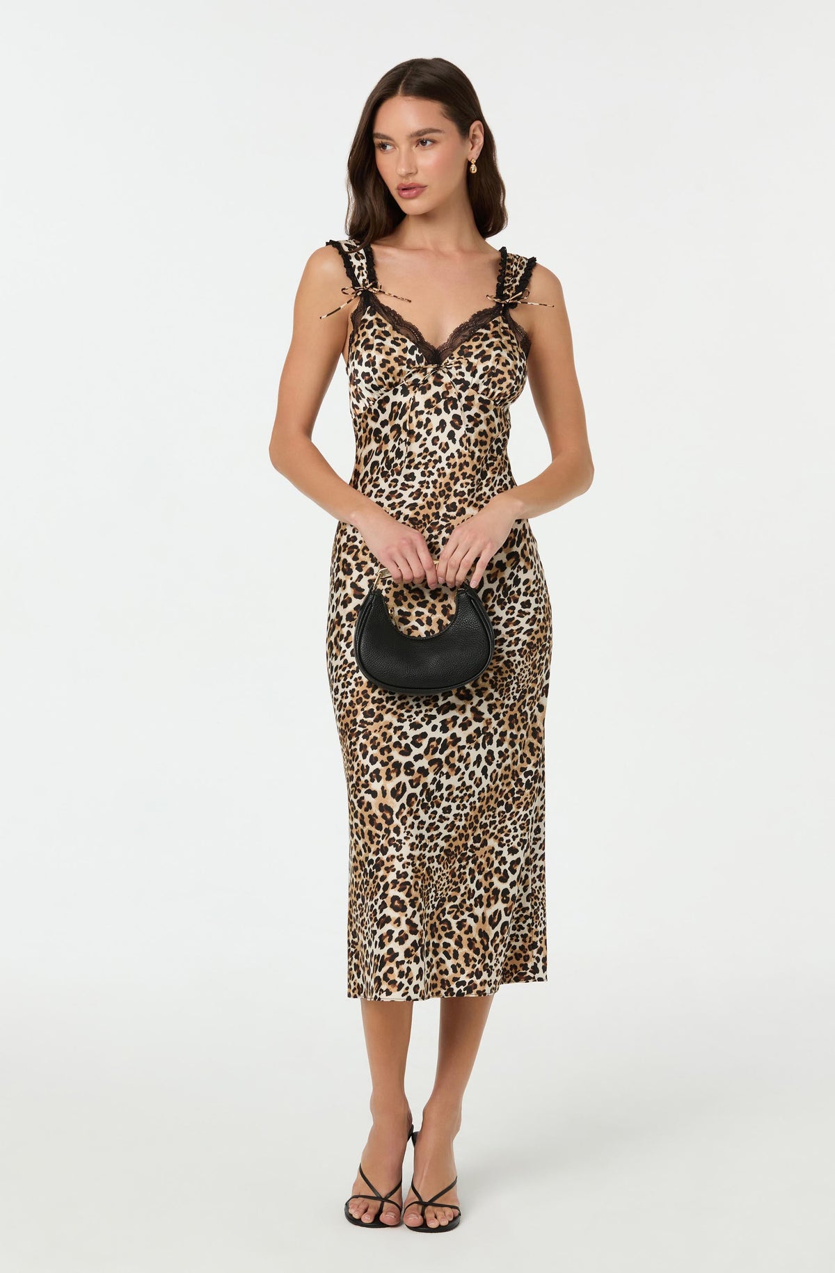 A woman is wearing a sleeveless, leopard print midi dress with black lace detailing on the shoulder straps and neckline. She is holding a small black handbag with a rounded shape and short handle, and is wearing black strappy high-heeled sandals.