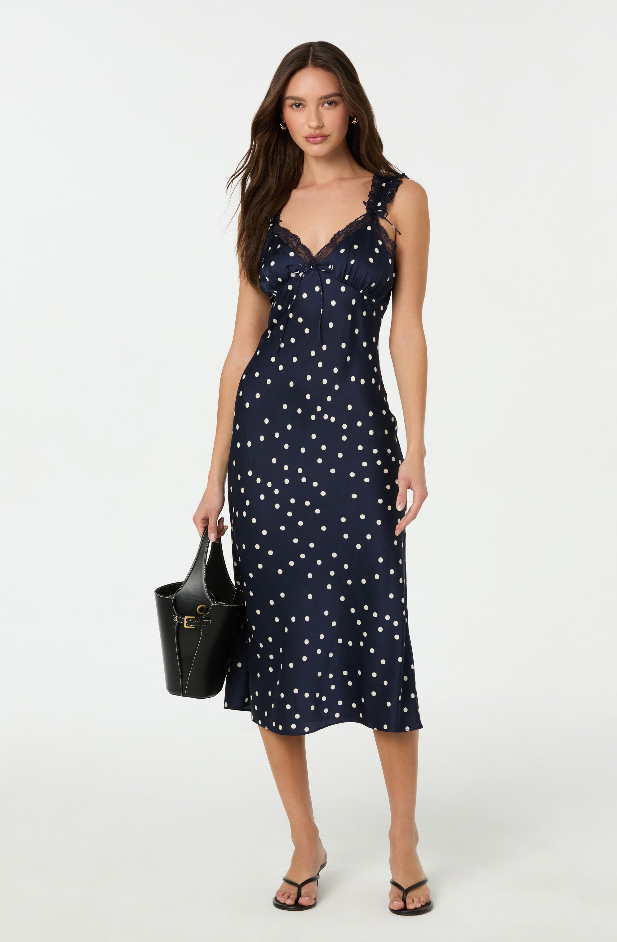 A woman is wearing a navy blue sleeveless dress with white polka dots, featuring lace details on the shoulder straps and neckline. She is carrying a black handbag with a gold clasp and is wearing black strappy sandals.