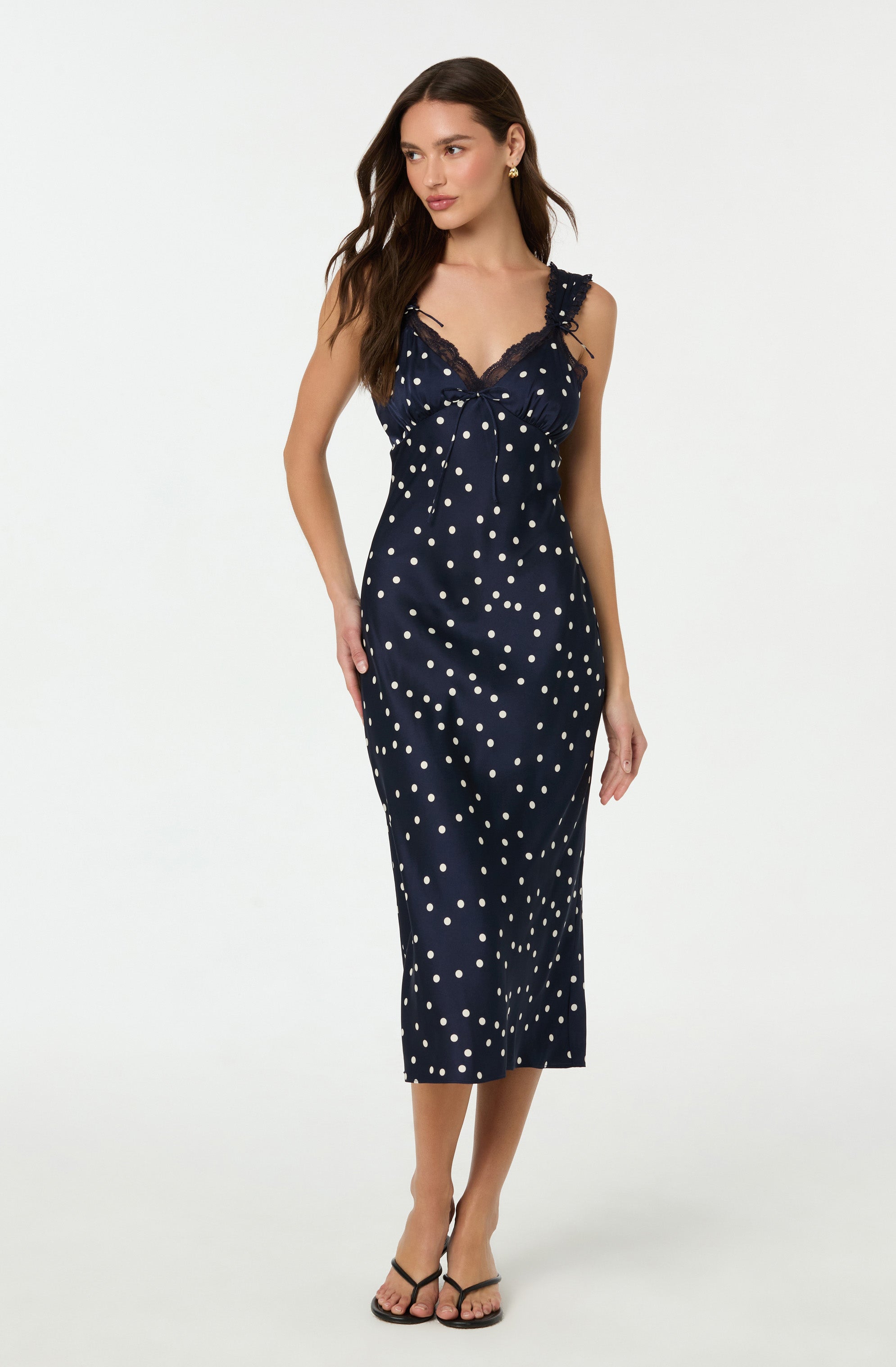 A woman is wearing a navy blue sleeveless slip dress with white polka dots. The dress features lace trim along the V-neckline and shoulder straps, and it falls to mid-calf length. She is also wearing black strappy high-heeled sandals.