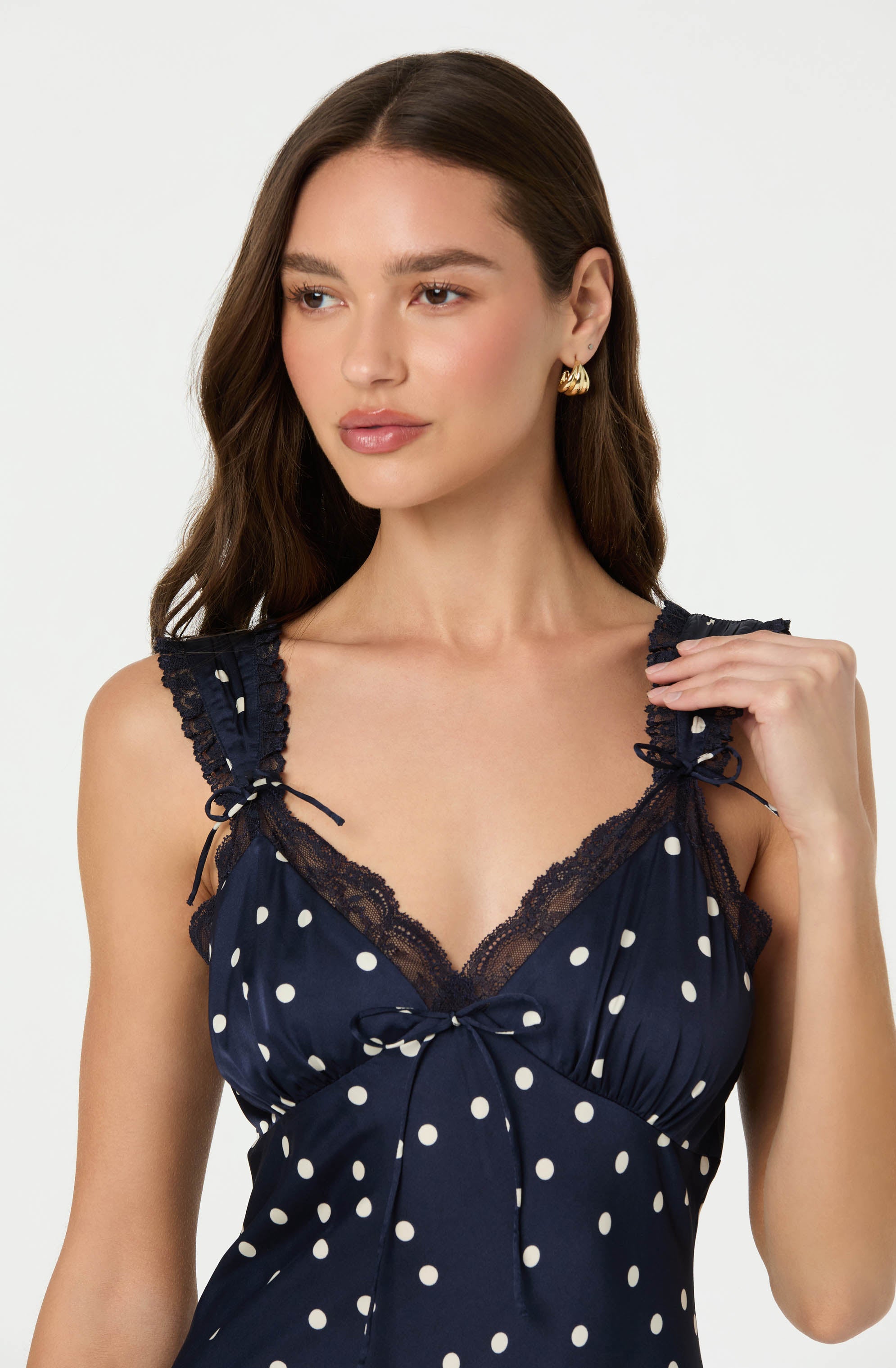 A woman is wearing a navy blue sleeveless top with white polka dots. The top has lace trim along the neckline and shoulder straps, which are tied into bows. The garment features a gathered bust area with a small bow at the center.