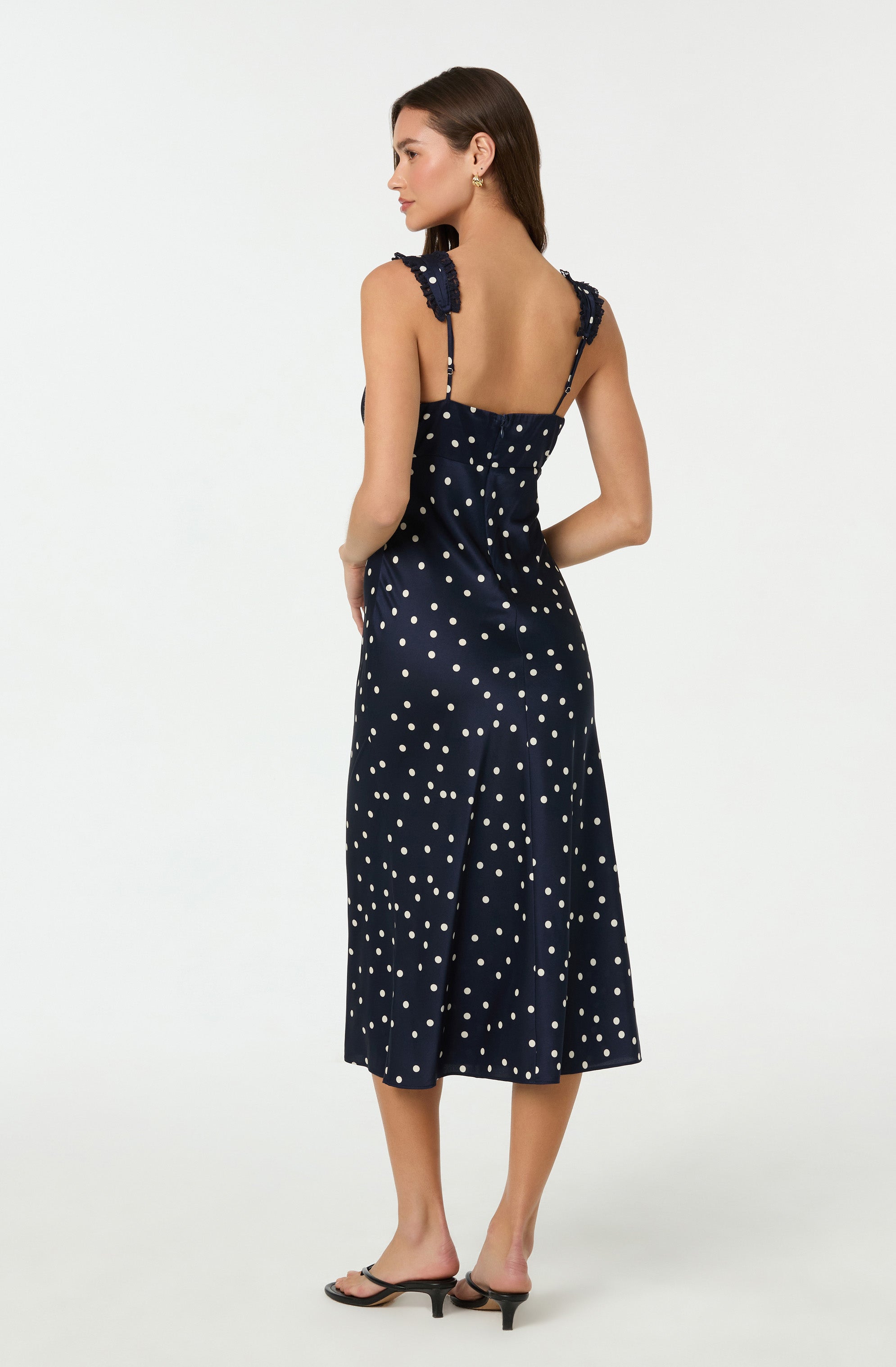 A woman is wearing a navy blue dress with white polka dots. The dress has wide shoulder straps with ruffle details and a fitted bodice that flares into a midi-length skirt. She is also wearing black open-toe heels.