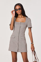 A woman is wearing a fitted, knee-length dress with a small checkered pattern in neutral tones. The dress has short sleeves, a square neckline, and a row of buttons down the front. It features two flap pockets at the hips. She is accessorizing with dark, narrow sunglasses, a gold watch, and a gold ring, and is holding a brown handbag.