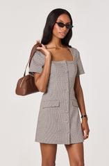 A woman wearing a short-sleeved, knee-length dress with a small checkered pattern in shades of beige and brown. The dress features a square neckline, a front row of buttons, and a pocket on each side at the hips. She is accessorizing with black sunglasses, a black wristwatch, a gold ring, and is carrying a brown handbag over her shoulder.