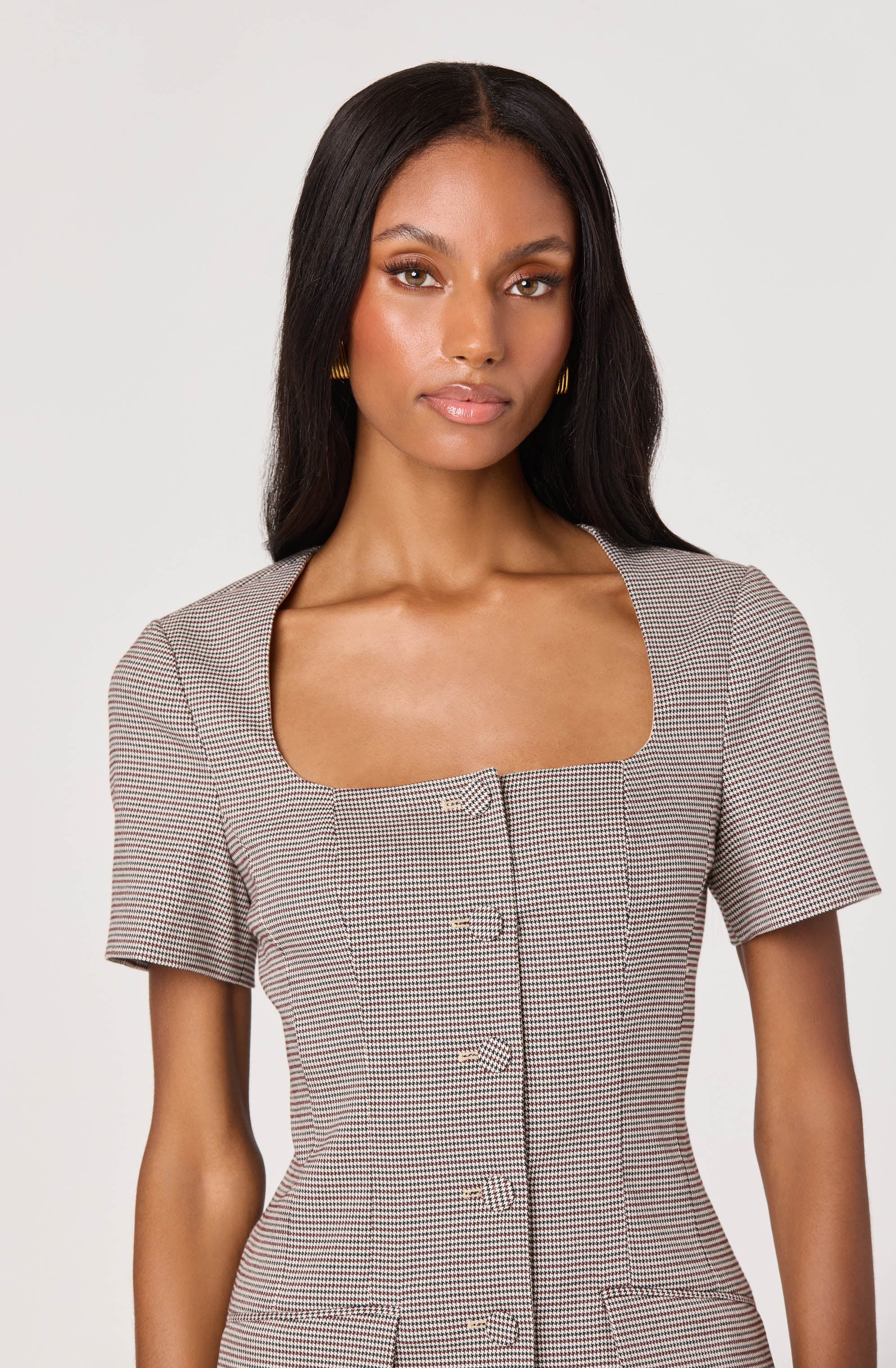 A woman with straight, dark hair is wearing a fitted dress with short sleeves, featuring a small checkered pattern in shades of black, white, and brown. The dress has a square neckline and a row of buttons down the front.