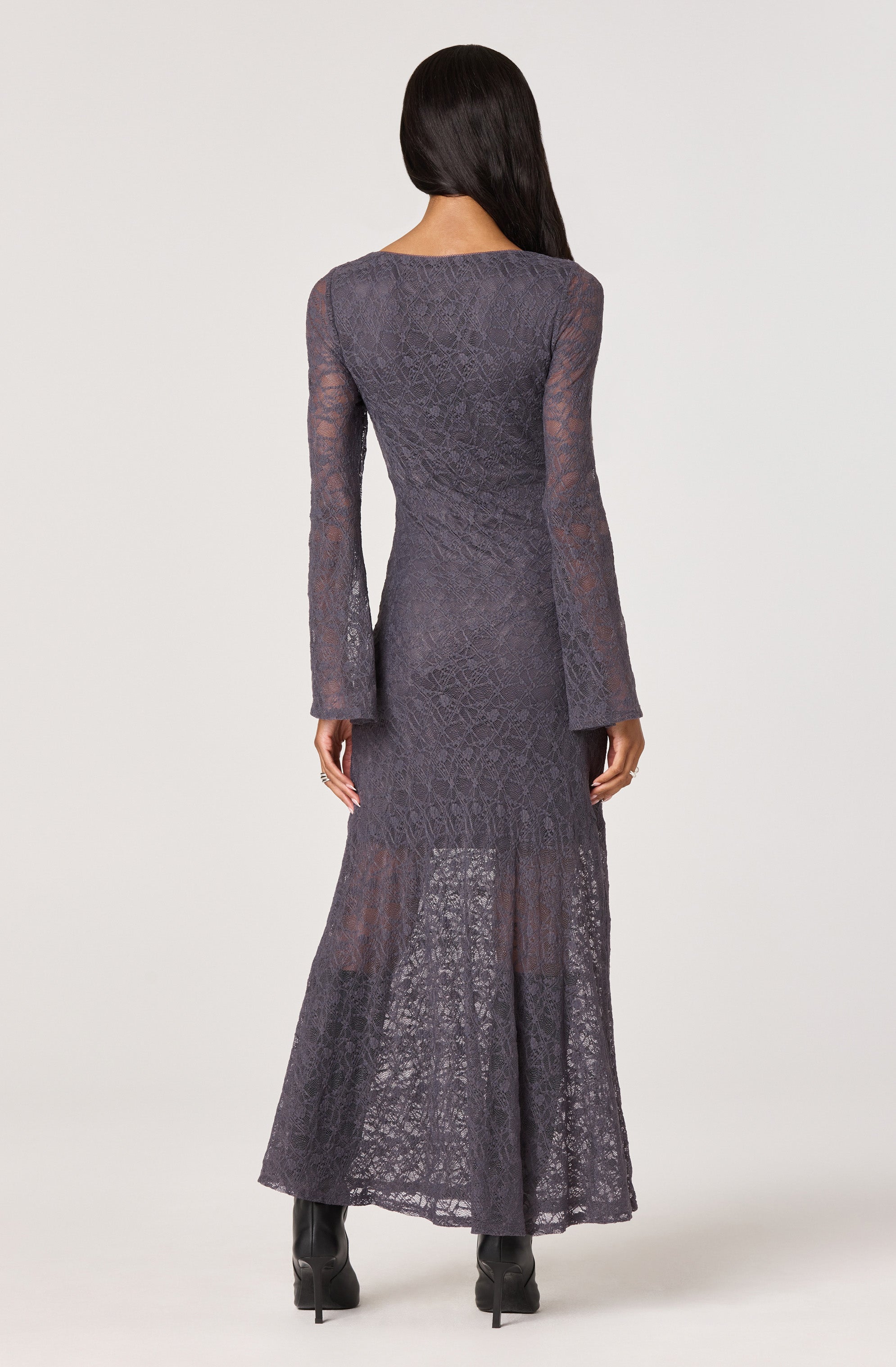 A woman is wearing a long, semi-sheer, dark gray lace dress with intricate patterns. The dress features long sleeves and a fitted bodice, extending down to a flared, slightly transparent skirt that reaches the floor.