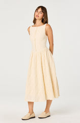 A woman is wearing a sleeveless, cream-colored, textured midi dress with a fitted bodice and a full, gathered skirt that falls below the knees. She is pairing the dress with light-colored loafers.