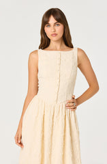 A woman wearing a sleeveless, cream-colored textured dress with a fitted bodice featuring a row of buttons down the front and a full, gathered skirt.