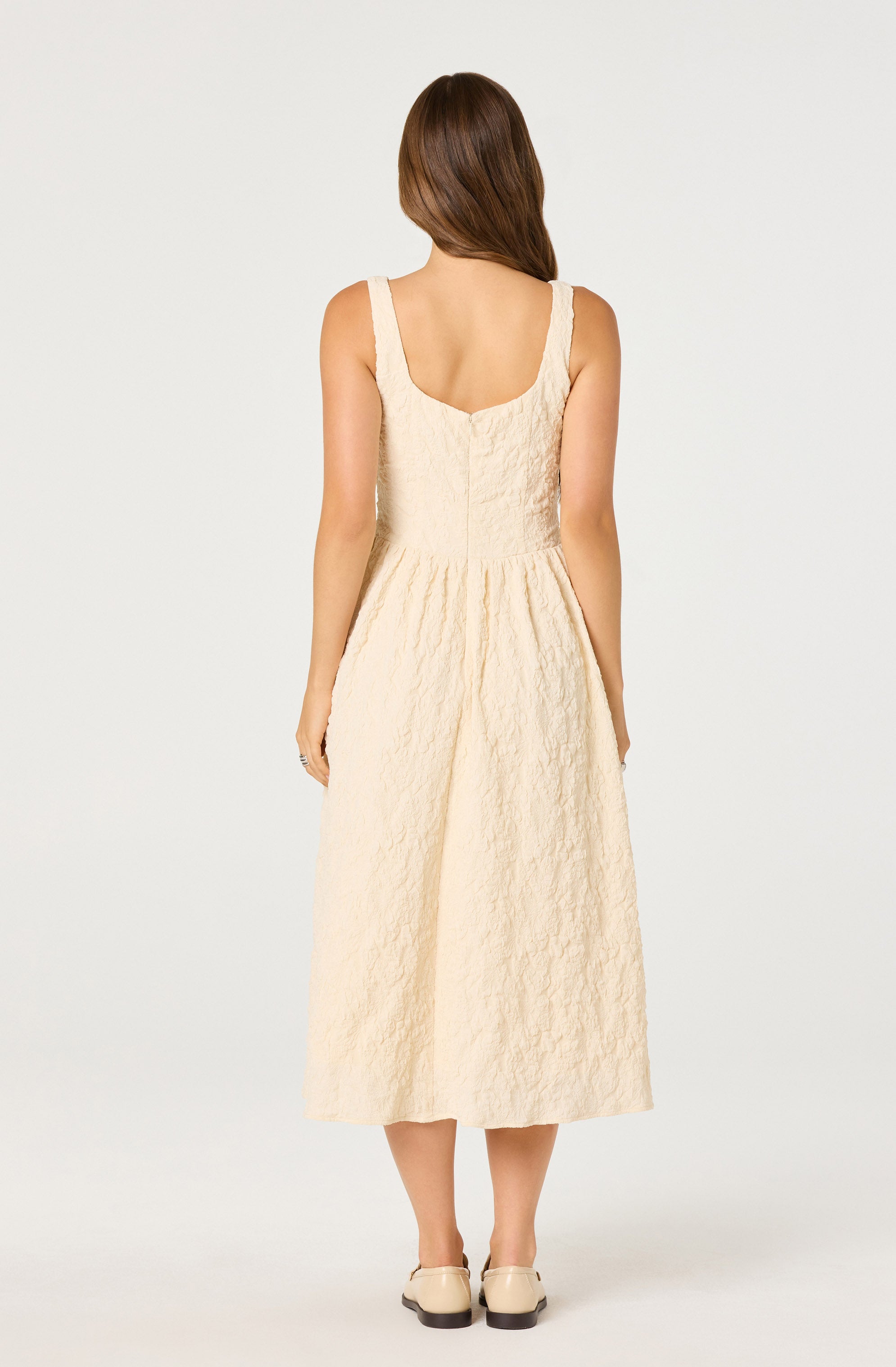A woman is wearing a sleeveless, ankle-length dress with a fitted bodice and a gathered waist. The dress is made of a textured, cream-colored fabric. She is also wearing beige, low-heeled shoes.