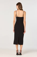 A woman is wearing a long, black sleeveless dress with thin adjustable spaghetti straps. The dress features a back zipper closure and a slit at the center bottom hem. She is also wearing black high-heeled sandals with ankle straps.