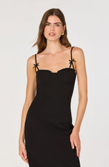 A woman is wearing a fitted, sleeveless black dress with thin spaghetti straps that are tied into small bows on her shoulders. The dress has a structured bust with a scalloped neckline edging. She is also wearing gold hoop earrings and a gold ring on her right hand.