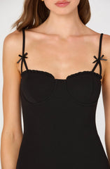 A woman is wearing a fitted black dress with thin spaghetti straps. The straps are decorated with small black bows on each side. The dress has a sweetheart neckline with a slight ruffle detail along the top edge. The fabric appears smooth and stretchy.
