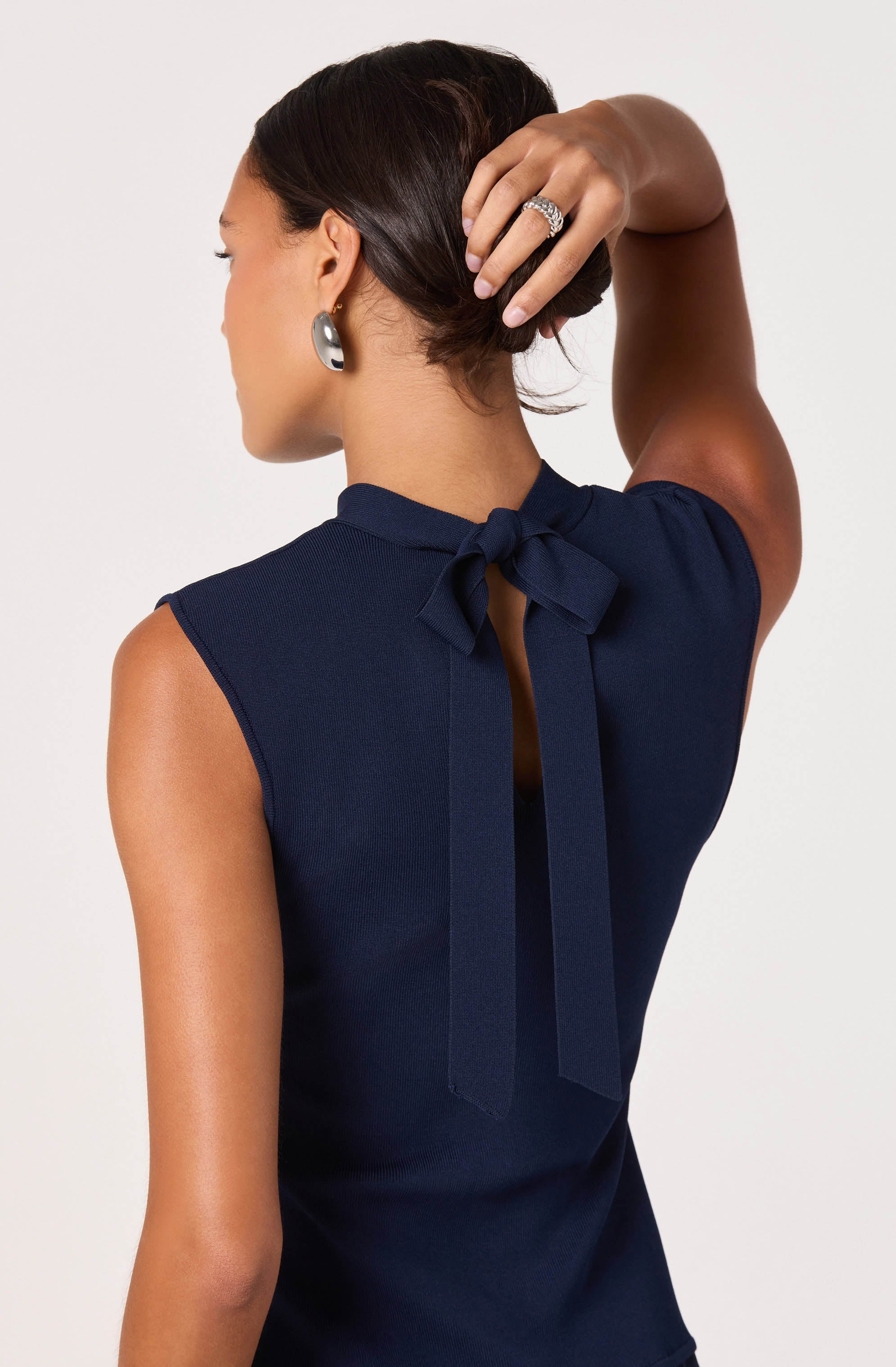 A woman is wearing a sleeveless navy blue top with a bow tied at the back of the neckline. The top has a small keyhole opening below the bow. She accessorizes with silver hoop earrings and a large ring on her right hand.