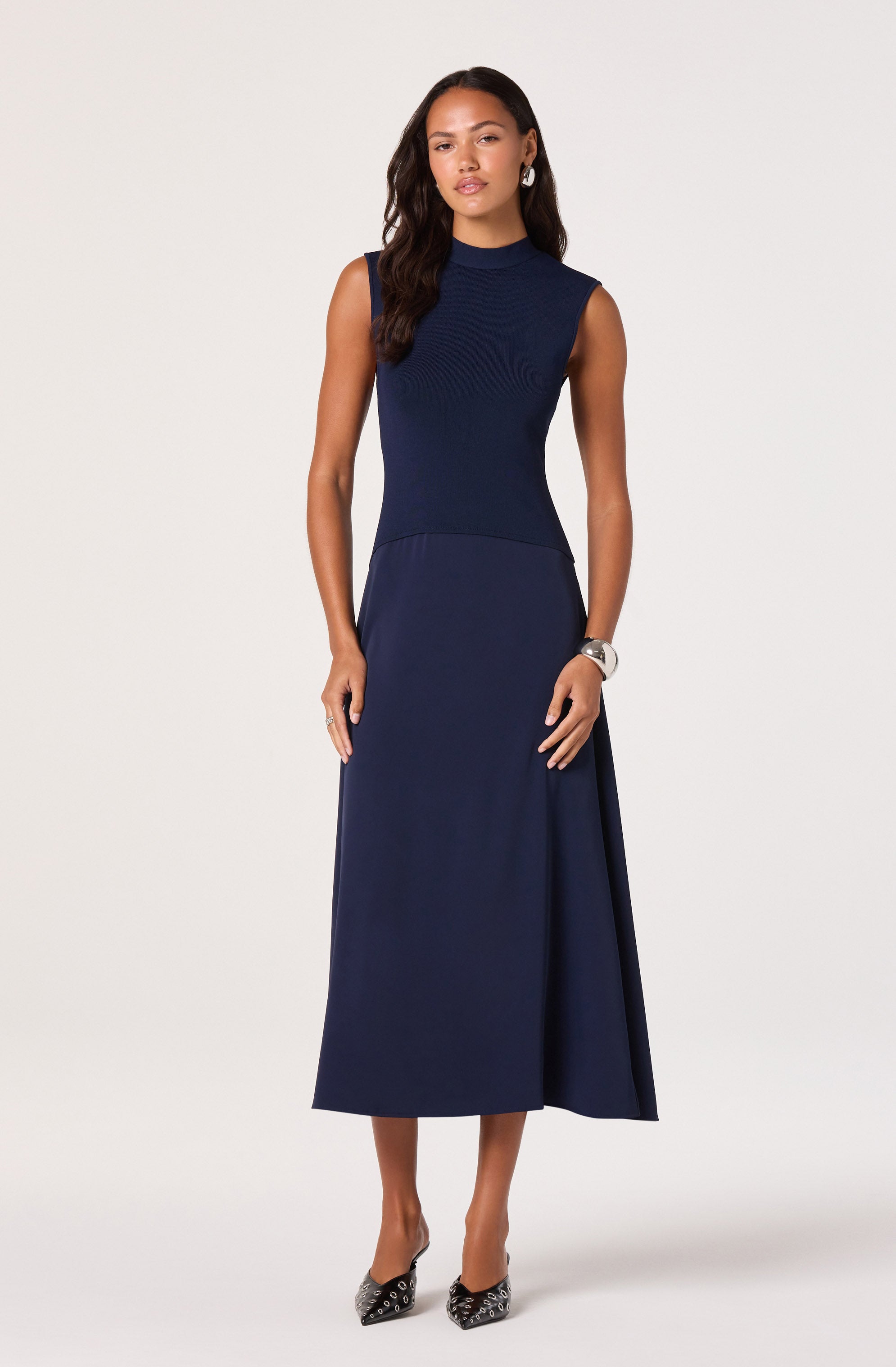 The woman is wearing a sleeveless, navy blue, high-neck dress that is fitted at the top and flares out into a long, flowing skirt. She has on patterned high heels and a wristwatch, with her hair styled in loose waves.