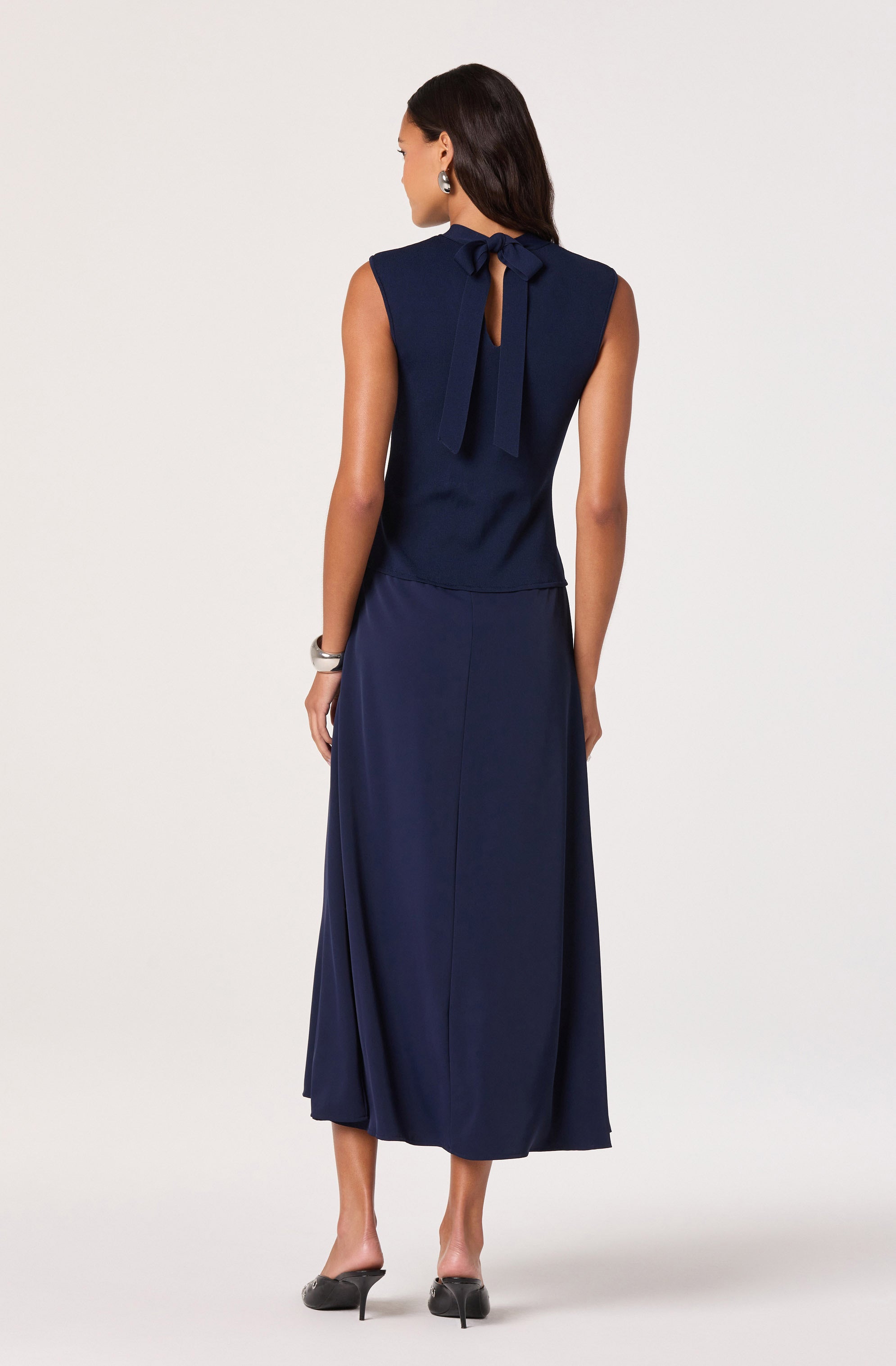 A woman is wearing a sleeveless navy blue dress with a fitted bodice and a long, flowing skirt. The dress features a keyhole opening at the back, tied with a large bow at the neckline. She accessorizes with pearl earrings and a wide bracelet on her right wrist. She is also wearing black high-heeled shoes.