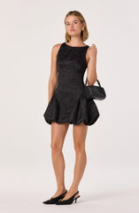 A young woman is wearing a sleeveless black dress with a fitted bodice and a voluminous, bubble-style skirt. The dress has a subtle textured pattern that adds visual interest. She completes her outfit with black pointed-toe heels and carries a small black handbag with a chain strap.