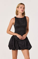A young woman is wearing a sleeveless black dress with a textured, subtle floral pattern. The dress has a fitted bodice and a voluminous, bubble-style skirt. She accessorizes with gold earrings and rings.