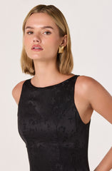 A woman is wearing a sleeveless black dress with a subtle floral pattern and a round neckline. She has paired the outfit with gold square-shaped earrings.