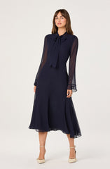 A woman is wearing a navy blue dress with sheer, long sleeves and a bow-tie collar. The dress is midi-length and flowy, with a slightly asymmetrical hemline. She is also wearing beige high-heeled shoes with ankle straps.
