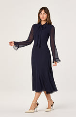 A woman is wearing a navy blue, midi-length dress with sheer, long sleeves and a bow tie at the collar. She pairs the dress with beige high-heeled shoes.