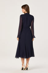 A woman is wearing a long, navy blue dress with sheer, long sleeves and a fitted bodice. The skirt is flowing and reaches mid-calf length. She pairs the dress with beige high-heeled sandals featuring ankle straps.