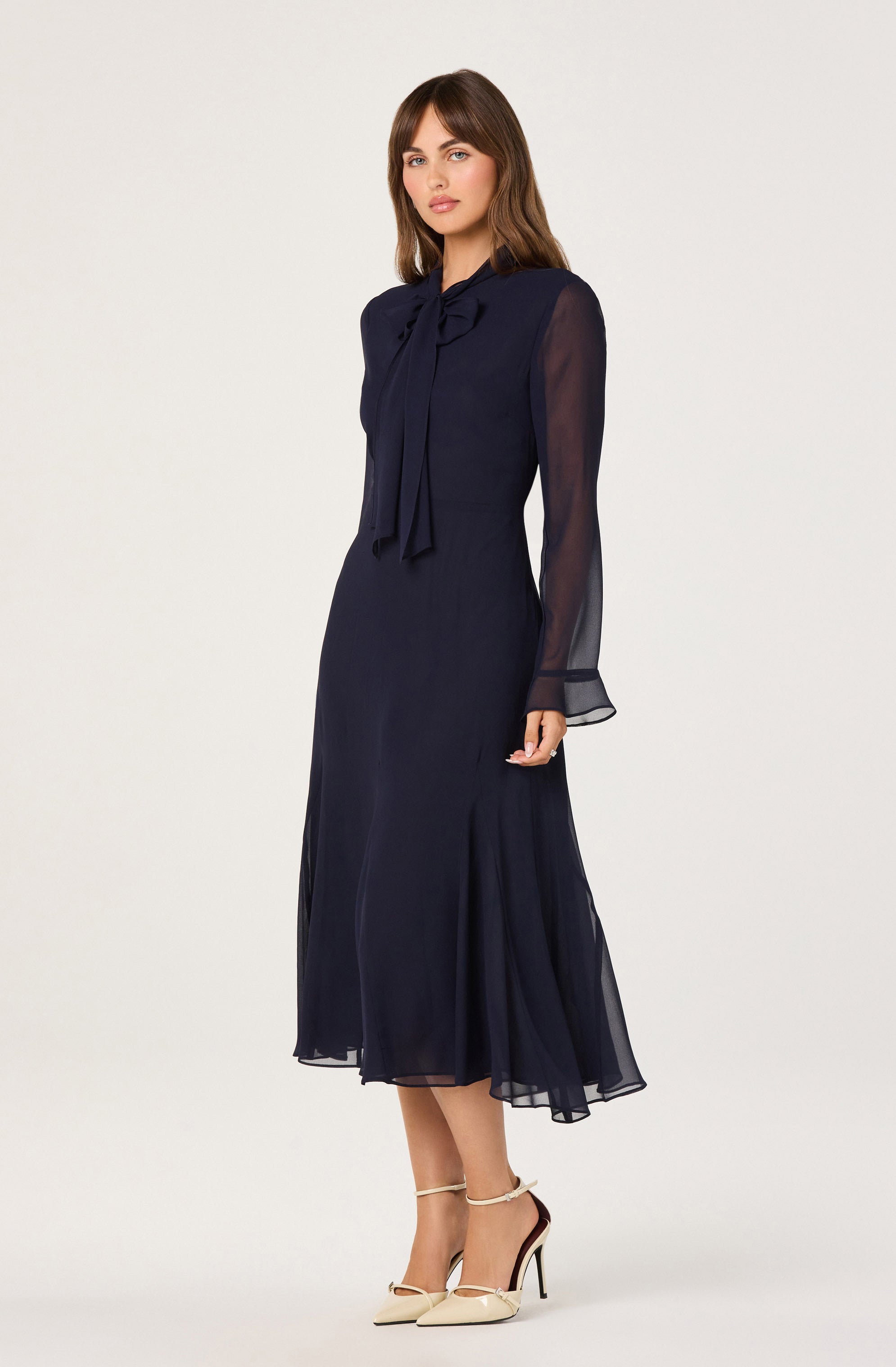 The woman is wearing a long, navy blue dress made of a sheer, flowing fabric. The dress features a high neckline with a large bow tie at the collar. The long sleeves are semi-transparent, with a slight puff at the shoulders and a flared, sheer cuff at the wrists.