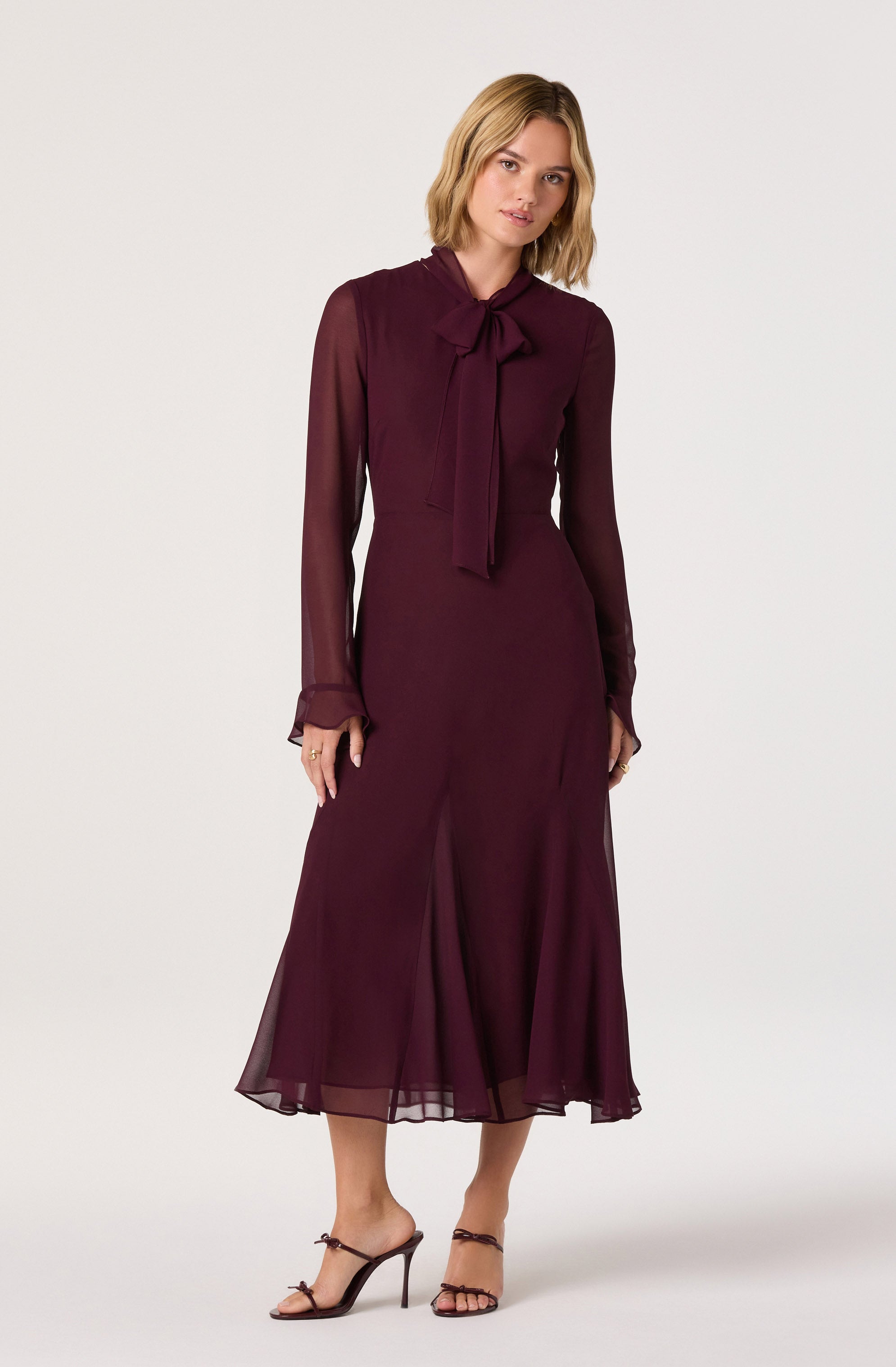 A woman is wearing a long, burgundy dress with sheer, long sleeves and a high neckline adorned with a bow tie. The dress has a fitted waist and a flowing, semi-transparent skirt that reaches mid-calf. She is also wearing black high-heeled sandals with thin straps.