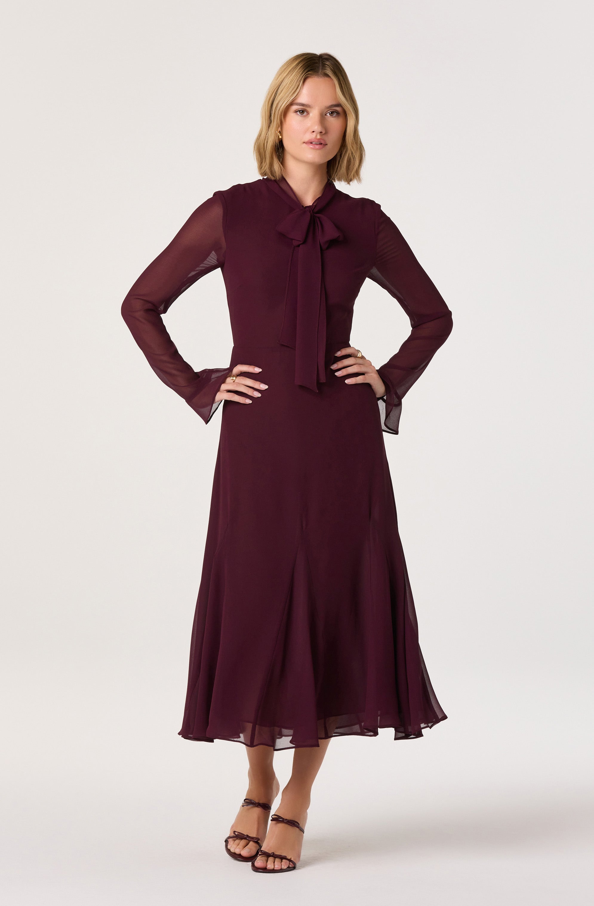 A woman is wearing a long, burgundy dress with sheer, long sleeves and a matching tie-neck bow. The dress has a fitted bodice and a flowing, floor-length skirt with vertical pleats. She is also wearing strappy high-heeled sandals.