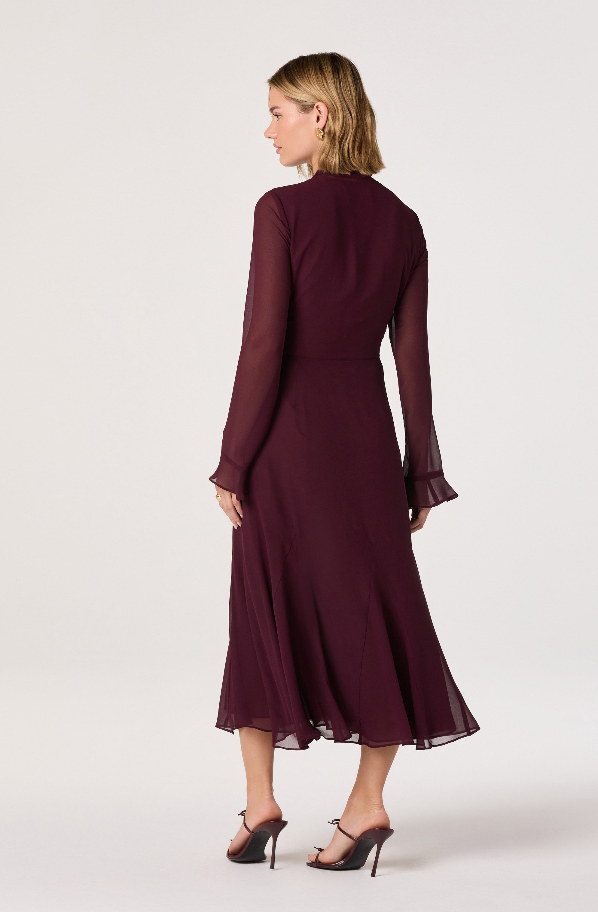 A woman is wearing a long, elegant dress in a deep burgundy color. The dress has sheer, long sleeves with ruffled cuffs and a fitted waist, flowing down to a midi-length skirt with a slight flare. She is also wearing high-heeled sandals with ankle straps.