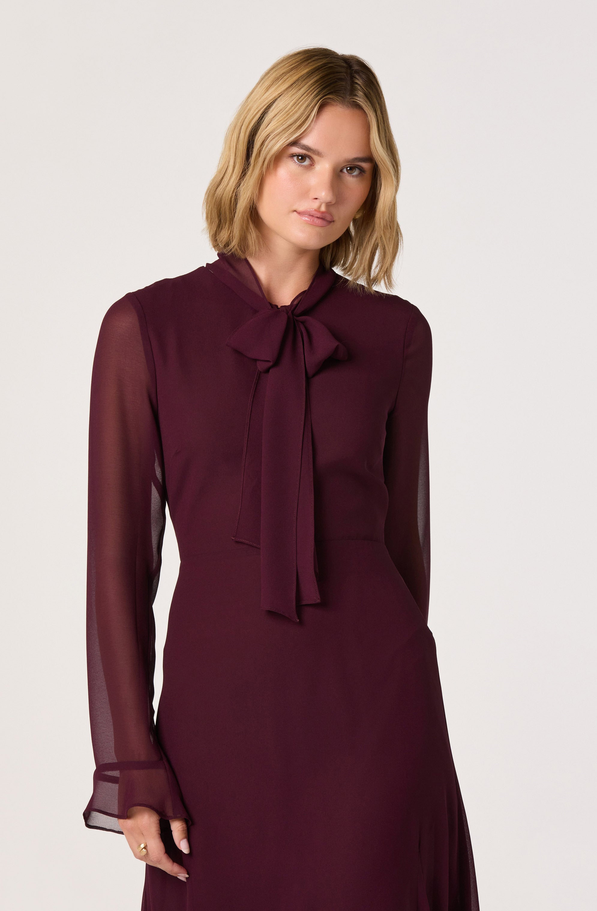 A woman is wearing a long-sleeved, deep burgundy dress with sheer sleeves that have a loose fit at the cuffs. The dress features a high neckline with a large bow tied at the neck. The fabric appears to be smooth and lightweight, with a fitted silhouette at the waist.