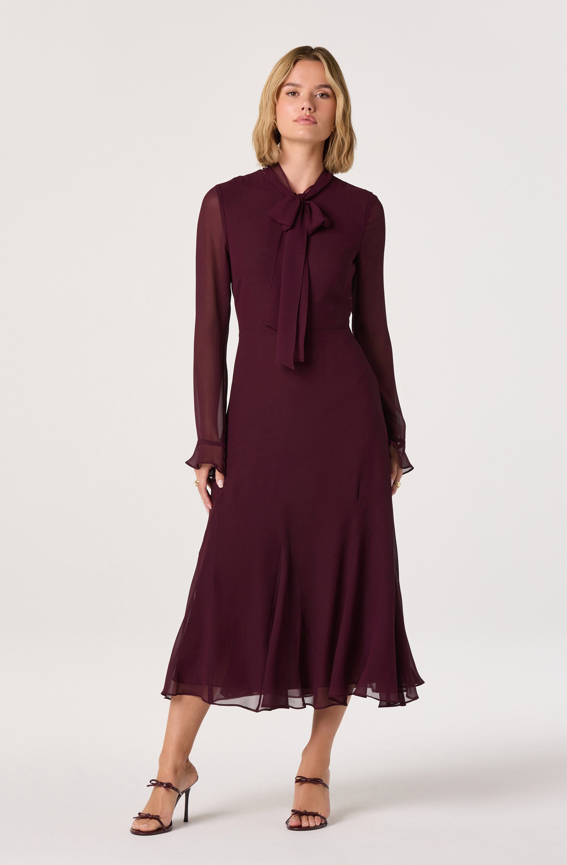 A woman is wearing a long, burgundy dress made of a sheer, flowy fabric. The dress features long, semi-transparent sleeves with ruffled cuffs and a large, matching bow tie at the collar. The skirt of the dress reaches mid-calf and has a slightly flared, uneven hem. She is also wearing strappy high-heeled sandals.