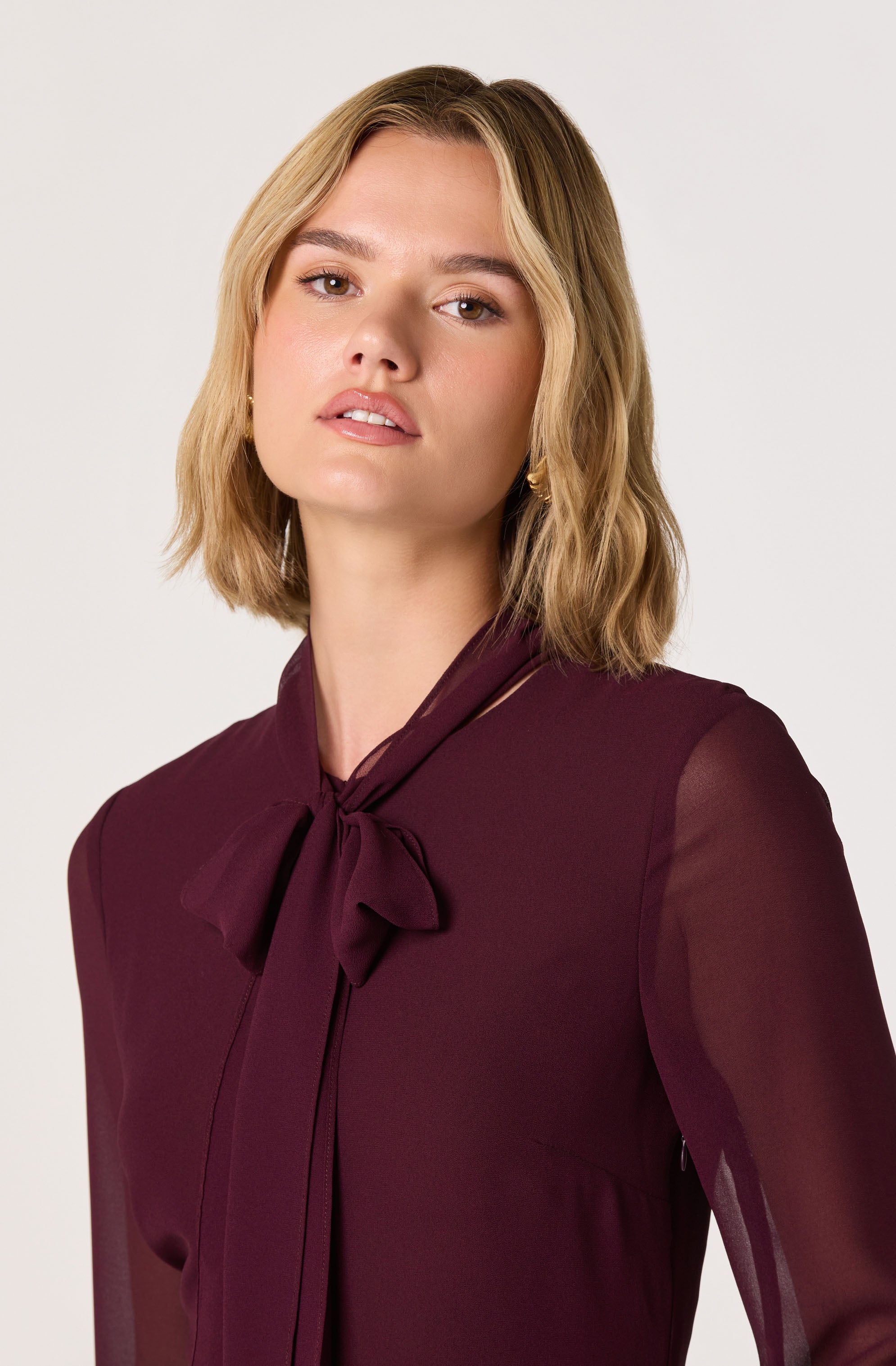 A woman with shoulder-length blonde hair is wearing a dark burgundy blouse with sheer long sleeves and a matching bow tie at the collar.
