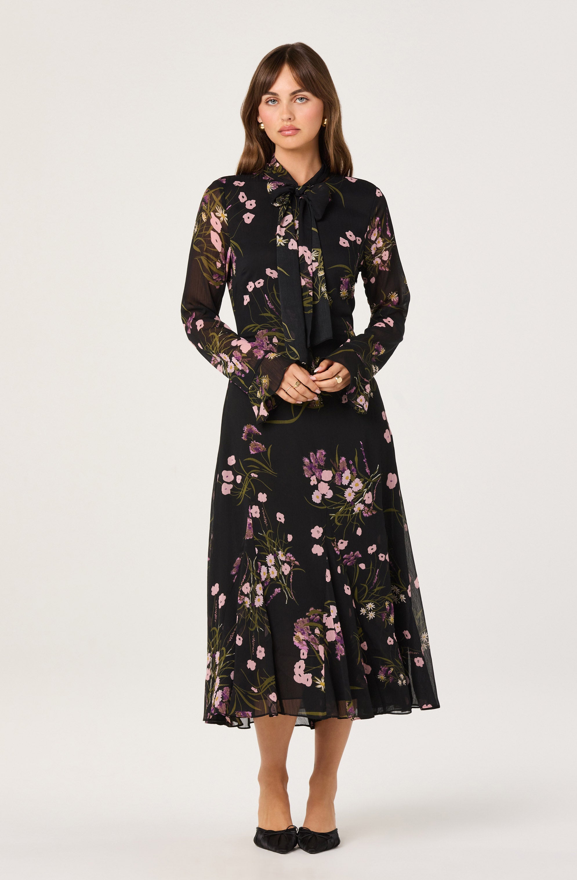 A woman is wearing a long, black dress with a floral pattern featuring pink, purple, and green hues. The dress has a sheer, lightweight fabric with a flowy silhouette and a midi length. She pairs the dress with a matching sheer blouse that has a large bow at the neckline, long sleeves, and a similar floral print. She completes her outfit with black pointed-toe shoes that have a bow detail.