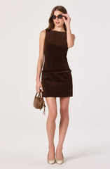 A woman is wearing a sleeveless, dark brown dress with a fitted top and a matching skirt that ends above the knees. She has on large, dark sunglasses and beige high-heeled shoes with a subtle pattern. She is holding a small beige handbag in her left hand.