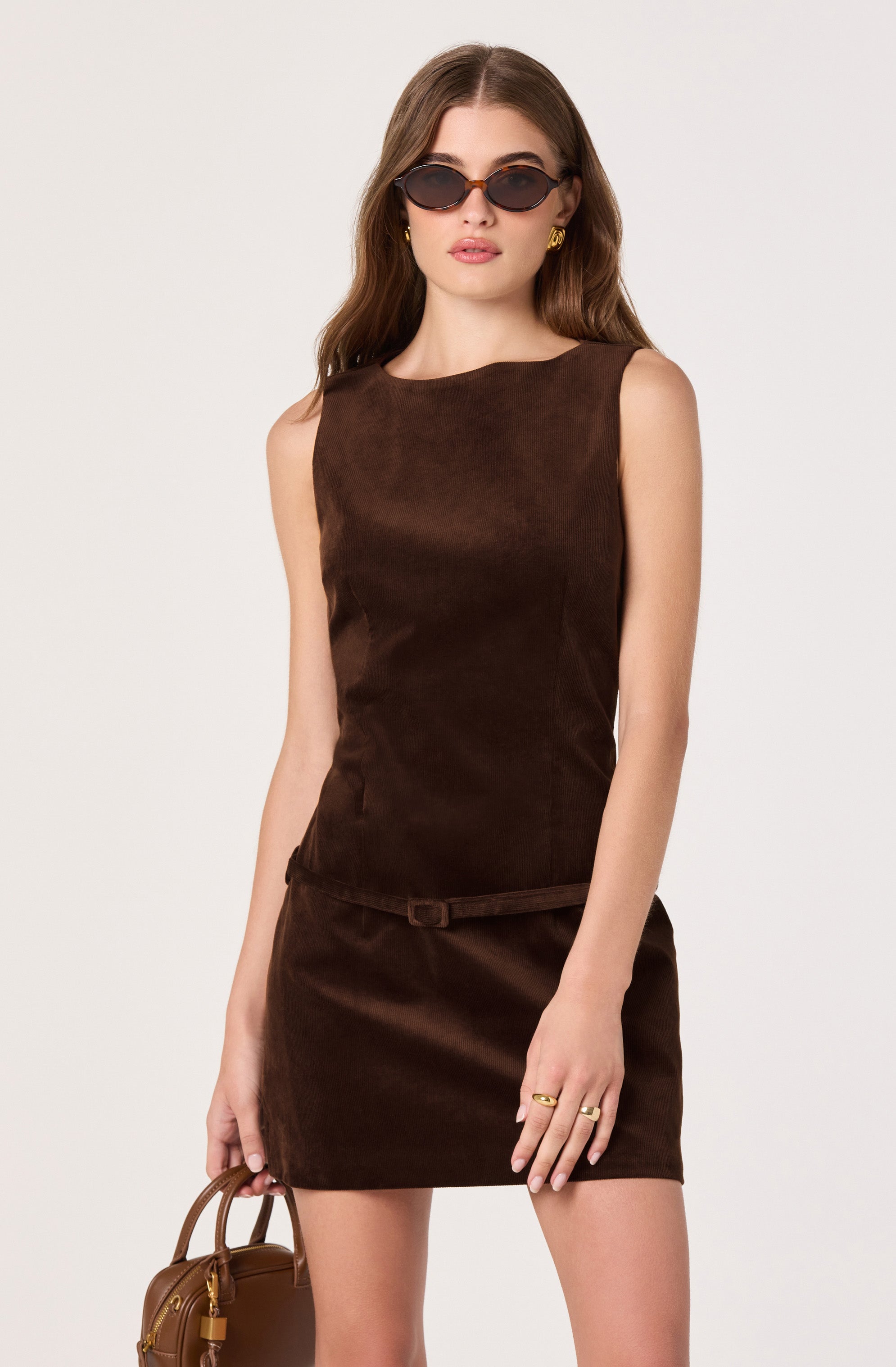 A woman is wearing a sleeveless, dark brown suede dress with a round neckline and a small matching belt at the waist. She accessorizes with black oval sunglasses, gold hoop earrings, and gold rings on her fingers. She is holding a small brown handbag in her left hand.