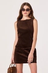 A woman is wearing a sleeveless, dark brown suede dress with a round neckline and a small matching belt at the waist. She accessorizes with black oval sunglasses, gold hoop earrings, and gold rings on her fingers. She is holding a small brown handbag in her left hand.
