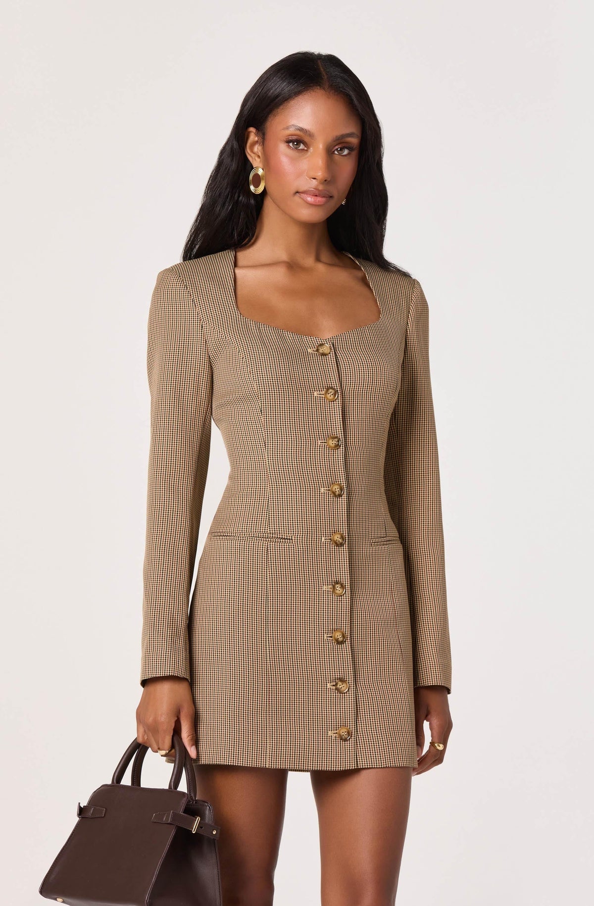 A woman is wearing a fitted, knee-length dress made of a light brown fabric with a small checkered pattern. The dress has long sleeves, a square neckline, and features a row of large, gold buttons down the front. She is holding a dark brown handbag in her right hand and has large, gold hoop earrings.