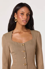 A woman is wearing a fitted, long-sleeved blazer with a small checkered pattern in shades of beige, brown, and black. The blazer features a square neckline and is fastened with large, shiny gold buttons down the front. She also has large, gold hoop earrings.