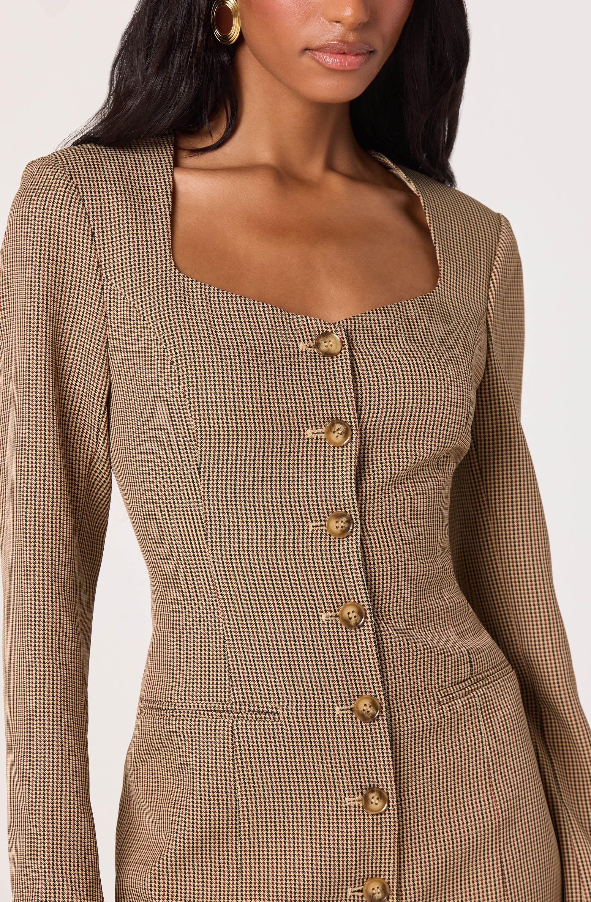 A woman is wearing a fitted blazer with a small checkered pattern in shades of brown, black, and beige. The blazer features a square neckline and is fastened with a row of large, round, gold buttons down the front. She is accessorized with large, gold hoop earrings.