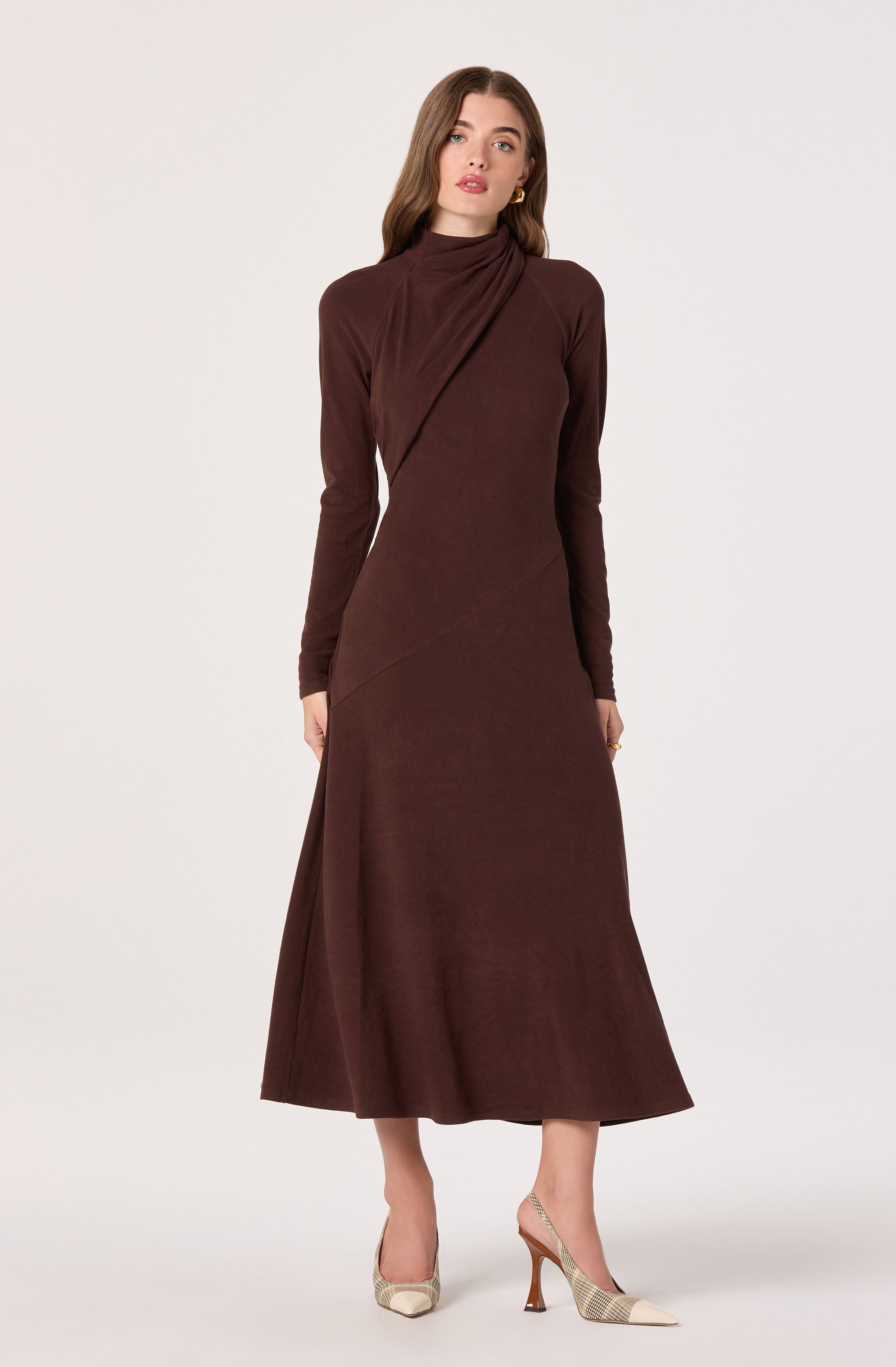 A woman is wearing a long, brown, high-neck dress with long sleeves. The dress has a slight sheen and features subtle diagonal seam details. She accessorizes with gold earrings and beige high-heeled shoes with a checkered pattern and an ankle strap.