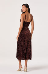 A woman is wearing a sleeveless, ankle-length dress with a fitted bodice and thin spaghetti straps. The dress has a rich, dark reddish-brown color with a floral pattern made of textured floral velvet fabric. She is accessorized with gold hoop earrings, a gold bracelet, and beige strappy high-heeled sandals.