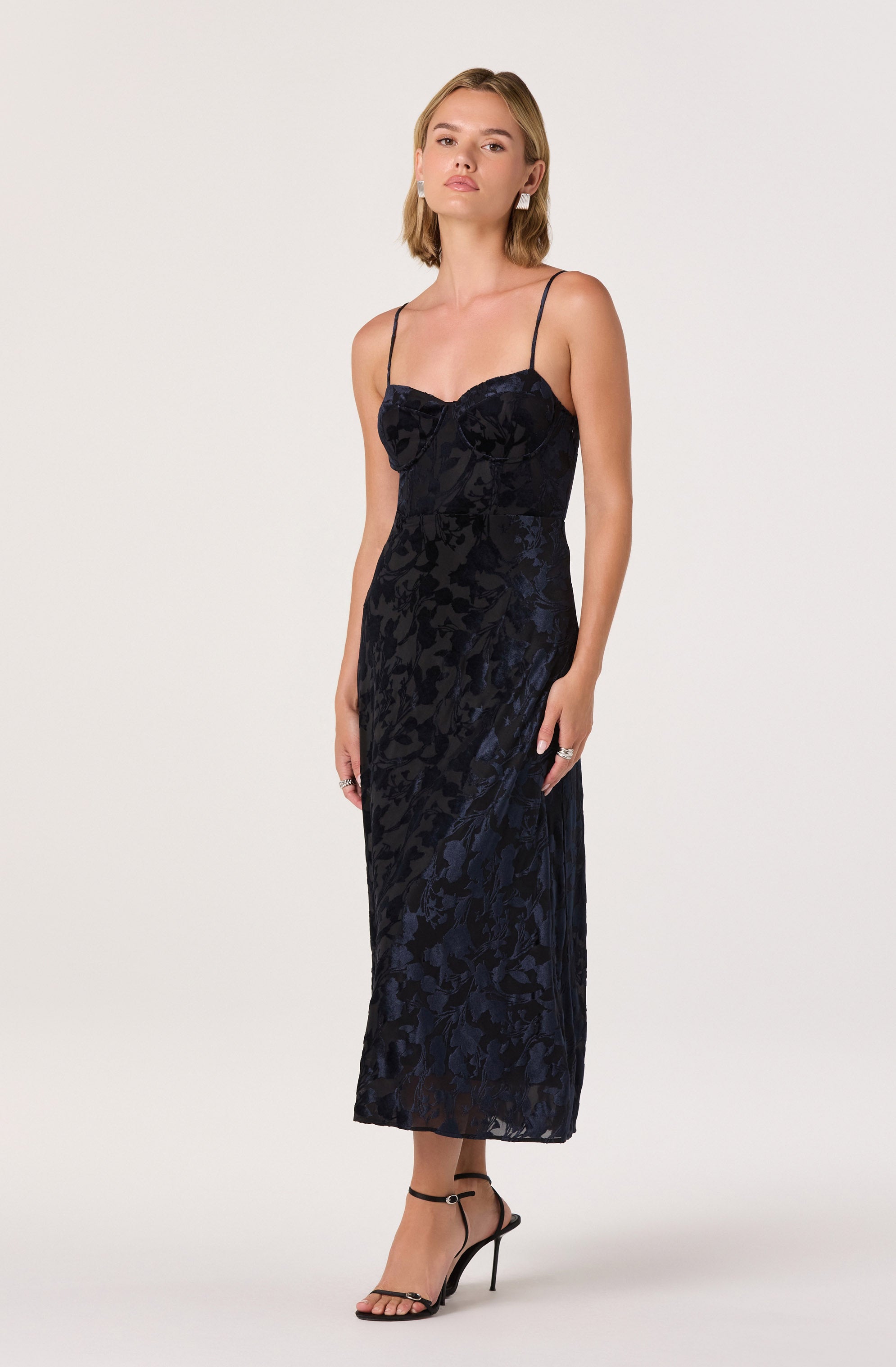 A woman is wearing a long, elegant black dress with a floral pattern. The dress has thin spaghetti straps and a fitted bodice with a bow detail at the center. She is also wearing strappy black high-heeled sandals.