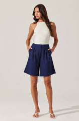 A woman is wearing a sleeveless, ribbed, white knit top with a high neckline. She pairs it with high-waisted, navy blue shorts featuring white pinstripes, pleats at the front, and side pockets. She is also wearing white strappy high-heeled sandals.