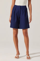 A person is wearing high-waisted navy blue shorts with thin white vertical pinstripes, pleats at the front, and a fabric belt tied at the waist. They are also wearing a sleeveless, white, ribbed top and white open-toe high-heeled sandals.