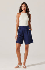 A woman is wearing a sleeveless, ribbed, cream-colored tank top and high-waisted, navy blue shorts with white pinstripes. She is also wearing white open-toe heels.