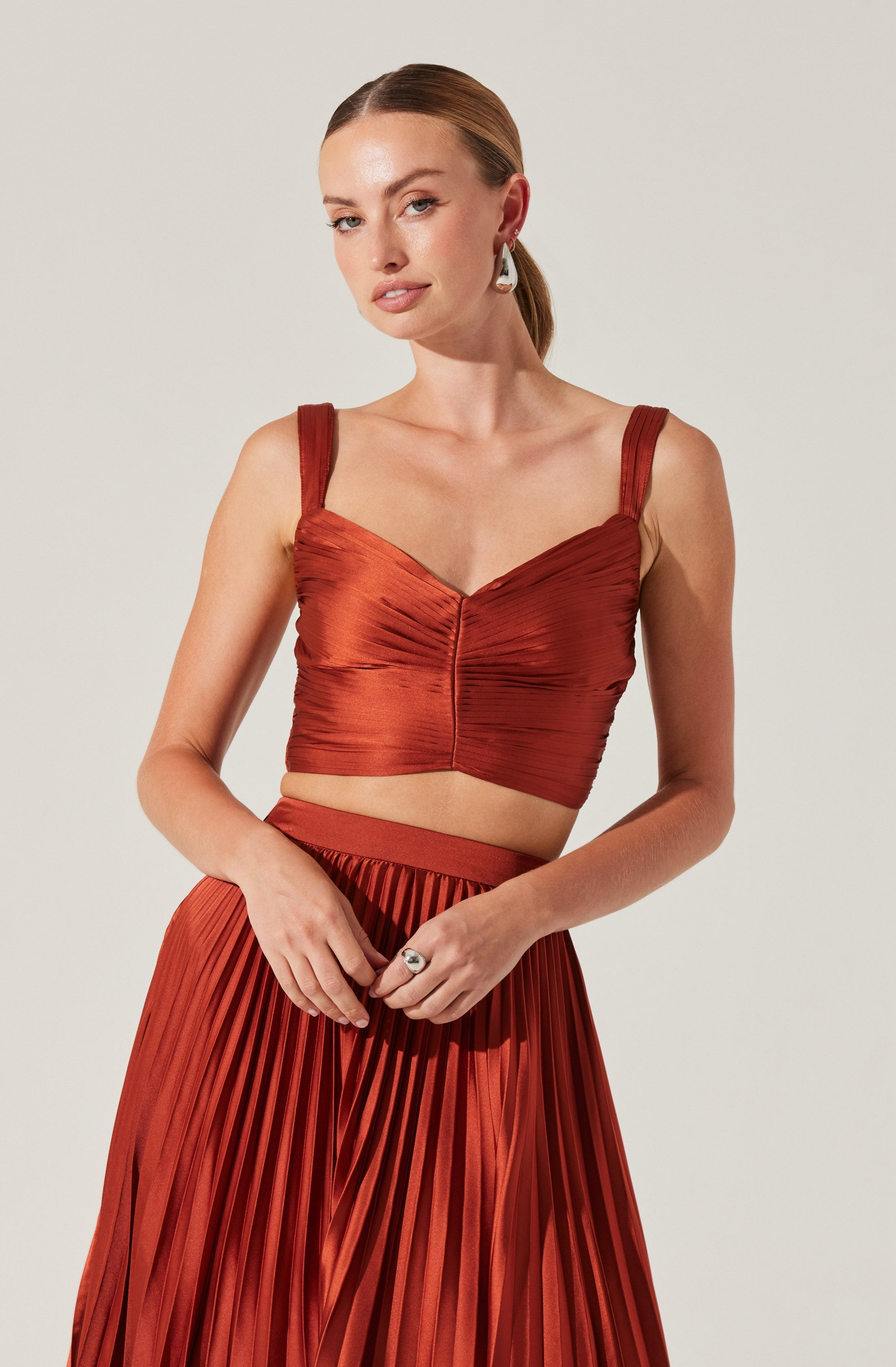 A woman is wearing a rust-colored, ruched crop top with wide shoulder straps and a sweetheart neckline. She is also dressed in a matching high-waisted, pleated skirt made of a shiny fabric.