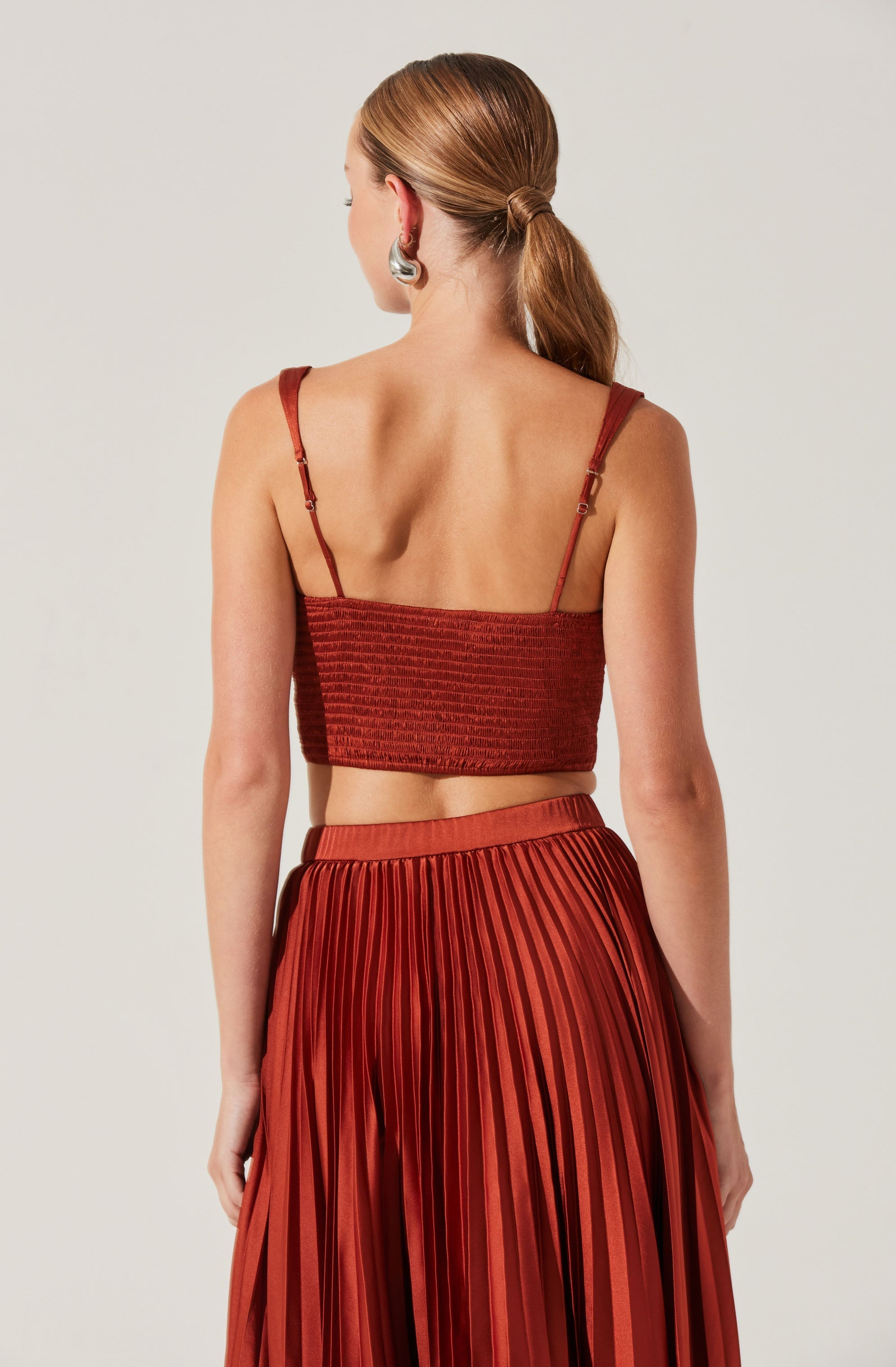 A woman is wearing a rust-colored, ribbed crop top with thin, adjustable spaghetti straps. She pairs it with a matching high-waisted, pleated skirt that features vertical pleats and a wide waistband.